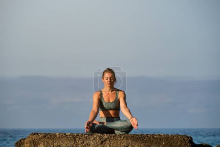 Téléchargez les photos : Corps complet de la jeune athlète féminine en vêtements de sport prenant Padmasana pose avec les mains dans le mudra gyan tout en étant assis sur le rocher et pratiquant le yoga parmi l'océan bleu - en image libre de droit