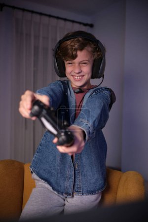 Téléchargez les photos : Souriant bouclé chevelu preteen kid dans des écouteurs et des vêtements décontractés debout sur un fauteuil tout en jouant jeu vidéo avec joystick - en image libre de droit