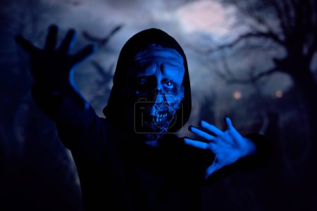 Foto de Zombie repugnante mirando a la cámara y haciendo gesto protector mientras está de pie bajo la luz de neón azul sobre el fondo borroso del cementerio oscuro - Imagen libre de derechos
