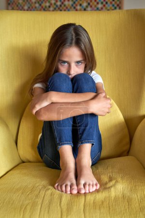 Foto de Cuerpo completo de niño descalzo infeliz mirando a la cámara mientras abraza las rodillas en un cómodo sillón en la sala de luz en casa - Imagen libre de derechos