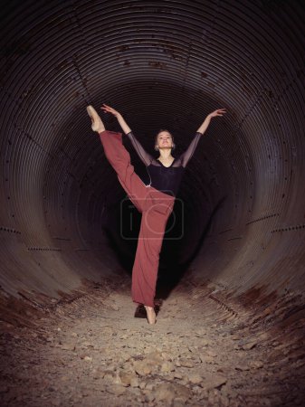 Téléchargez les photos : Corps complet de danseuse de ballet en chemisier noir et pantalon rouge effectuant un mouvement de danse gracieux avec les bras levés et la jambe contre le passage minable - en image libre de droit