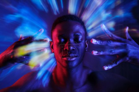 Téléchargez les photos : Calme jeune homme afro-américain avec torse nu tenant les mains près du visage tout en se tenant les yeux fermés dans des lumières bleues floues en studio sombre - en image libre de droit