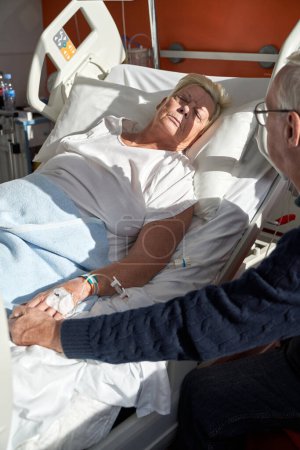Foto de De arriba de la cosecha el hombre mayor de la mano de la esposa enferma acostado en el sofá médico con catéter venoso conectado durante el proceso de rehabilitación en la clínica moderna - Imagen libre de derechos