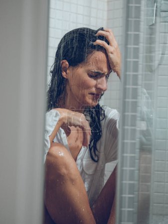 Téléchargez les photos : Femme triste avec de longs cheveux obtenir douche en chemise blanche tout en étant assis derrière un rideau transparent - en image libre de droit