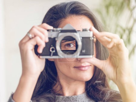 Photo for Smiling female with long dark hair looking through hole for lens in retro film photo camera while covering face against blurred background - Royalty Free Image