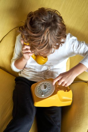 Photo for From above of boy talking on old fashioned wired telephone while sitting in comfortable armchair in light living room at home - Royalty Free Image
