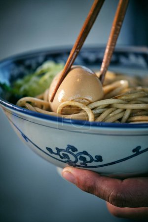 Photo for Crop anonymous person with boiled egg between food sticks in bowl with delicious Japanese noodle soup - Royalty Free Image