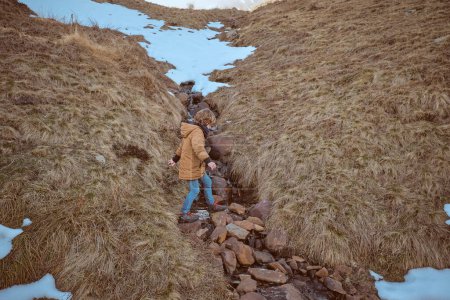Téléchargez les photos : D'en haut d'un garçon anonyme marchant sur un ruisseau avec des pierres sur un rivage herbeux avec de la neige dans la nature par temps froid - en image libre de droit