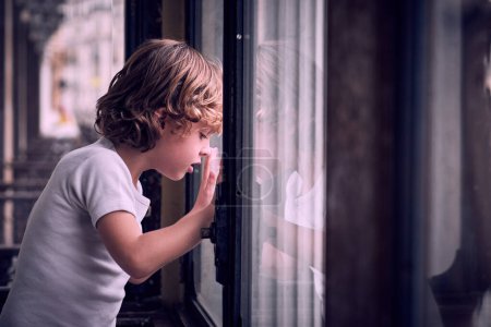 Téléchargez les photos : Vue latérale d'un petit garçon attentif aux cheveux blonds bouclés en t-shirt blanc regardant par la fenêtre avec curiosité à la maison en plein jour - en image libre de droit