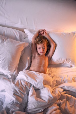 Photo for Top view of cute shirtless boy with raised arms lying under duvet in soft bed and sleeping in evening at home - Royalty Free Image