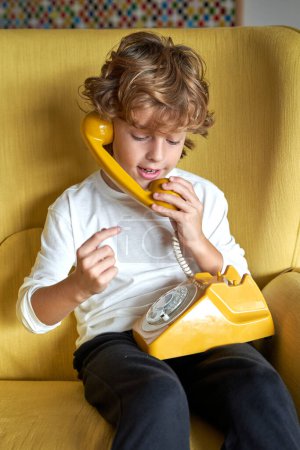 Téléchargez les photos : Preteen enfant avec les cheveux bouclés parlant sur le téléphone jaune rétro tout en étant assis dans un fauteuil confortable dans le salon à la maison - en image libre de droit