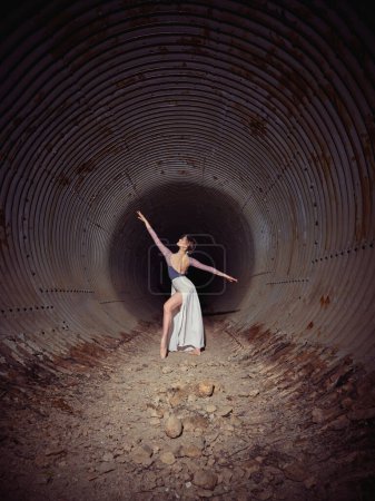 Téléchargez les photos : Vue latérale complète du corps d'une danseuse de ballet mince regardant le bras levé tout en dansant dans un vieux tunnel avec des pierres - en image libre de droit