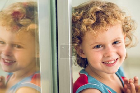 Photo for Smiling adorable boy with curly blond hair looking away while sitting near window with reflection on balcony of residential apartment - Royalty Free Image
