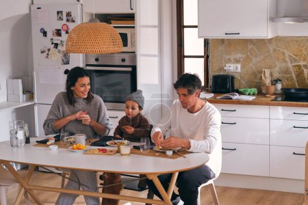 Téléchargez les photos : Femme souriante contre fille et mari à table avec délicieux petit déjeuner dans la cuisine de la maison - en image libre de droit