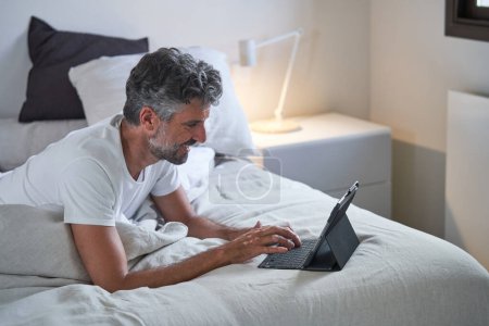 Photo for Side view of smiling middle aged male freelancer with beard and curly hair using tablet and typing on docking station while lying on bed and looking at screen - Royalty Free Image