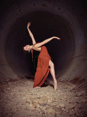 Foto de Vista lateral de cuerpo completo de talentosa bailarina de ballet en vestido doblando la espalda y levantando el brazo mientras baila en viejo túnel - Imagen libre de derechos