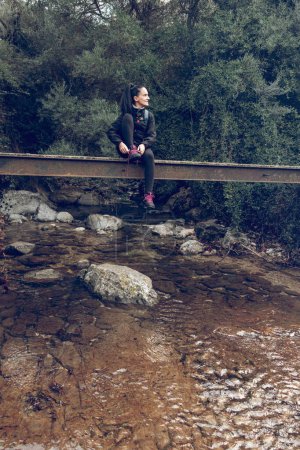 Téléchargez les photos : Touriste féminine en vêtements décontractés assis sur un pont de pierre au-dessus de la rivière dans la forêt de Majorque et regardant loin - en image libre de droit