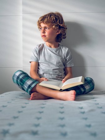 Foto de Lindo colegial de pelo rizado sentado en la cama con las piernas cruzadas con libro abierto y mirando hacia otro lado soñadoramente - Imagen libre de derechos