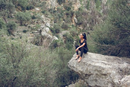 Téléchargez les photos : Heureuse femme pieds nus en vêtements noirs souriant et observant la chaîne de montagnes tout en étant assise sur un rocher dans la vallée de Majorque, Espagne - en image libre de droit