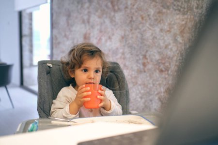 Photo for Adorable toddler sitting in chair with cup of hot drink and looking away in light room at home - Royalty Free Image