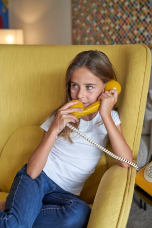 Photo for Child sitting in armchair in living room and talking on vintage rotary dial telephone while looking away at home - Royalty Free Image