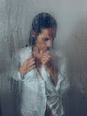 Téléchargez les photos : Grâce à un verre de femme triste en chemisier humide regardant vers le bas dans la cabine de douche près humide porte transparente dans la salle de bain légère - en image libre de droit