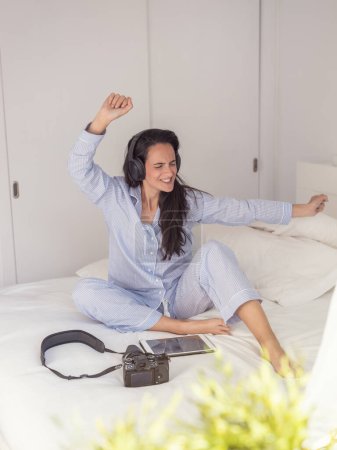 Foto de Mujer satisfecha en auriculares con ropa de dormir y bailando con los ojos cerrados en la cama mientras disfruta de la música en auriculares - Imagen libre de derechos