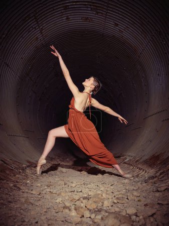 Photo for Full body side view of slim ballerina in red dress and pointe shoes dancing ballet in old tunnel with shabby walls - Royalty Free Image
