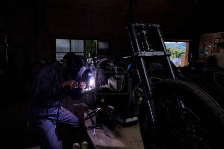 Photo for Anonymous male worker in uniform and shield using welding machine to fix some metal bike parts while working in dark workshop - Royalty Free Image