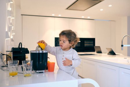 Téléchargez les photos : Adorable petit enfant en tenue décontractée préparant un délicieux jus d'orange pour un petit déjeuner sain dans la cuisine domestique à la maison - en image libre de droit