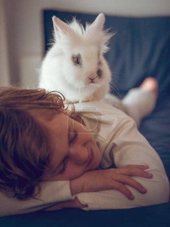 Foto de Lindo chico en pijama durmiendo tranquilamente en la cama cómoda con conejo blanco domesticado en la espalda en el dormitorio de luz en casa - Imagen libre de derechos