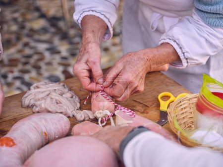 Photo for High angle of crop unrecognizable senior person in uniform using threads while stanching fresh raw stuffed sausages standing at wooden table in traditional Spanish butchery - Royalty Free Image