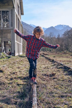 Foto de Cuerpo completo de niño en ropa casual balanceándose en el ferrocarril que pasa por el valle abandonado herboso rural rodeado de montañas - Imagen libre de derechos