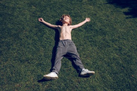 Téléchargez les photos : D'en haut du petit enfant insouciant avec le torse nu et les cheveux blonds bouclés couchés sur la pelouse verte avec les bras levés et les yeux fermés dans la journée ensoleillée d'été - en image libre de droit
