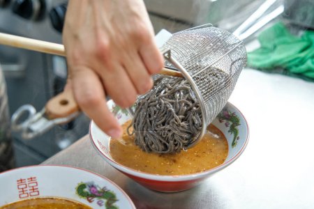 Photo for High angle of crop anonymous male chef adding soba noodles from stainless strainer basket in ceramic bowl of spicy soup in Asian food restaurant - Royalty Free Image