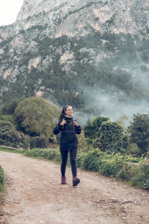 Téléchargez les photos : Touriste féminine pleine longueur avec sac à dos marchant seul sur le sentier près de la montagne verte et regardant loin dans la lumière du jour à Majorque - en image libre de droit