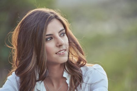 Photo for Attractive female with long dark wavy hair looking into distance while sitting in nature against blurred background on summer day - Royalty Free Image