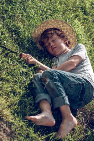Foto de Desde arriba de cuerpo completo lindo descalzo preadolescente niño en sombrero de mimbre y jeans acostado sobre hierba verde y quedarse dormido a la luz del sol - Imagen libre de derechos