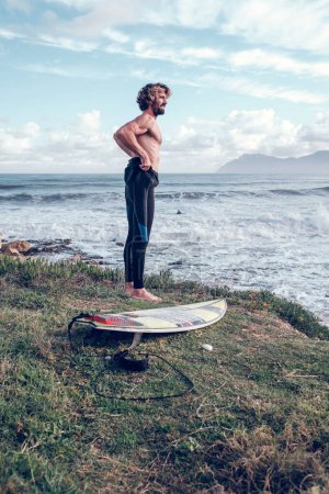 Foto de Vista lateral de un joven surfista masculino hispano musculoso poniéndose un traje de neopreno mientras está de pie en la costa cubierta de hierba cerca de la tabla antes de practicar surf en el océano ondulado - Imagen libre de derechos