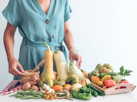 Photo for Crop unrecognizable female demonstrating assorted ripe fresh vegetables including pumpkins and tomatoes with pepper placed in wooden tray on table against white background - Royalty Free Image