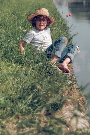 Téléchargez les photos : Corps complet d'enfant heureux pieds nus en chapeau de paille et jeans reposant sur le rivage herbeux vert du canal calme tout en profitant de la journée d'été et en regardant la caméra - en image libre de droit