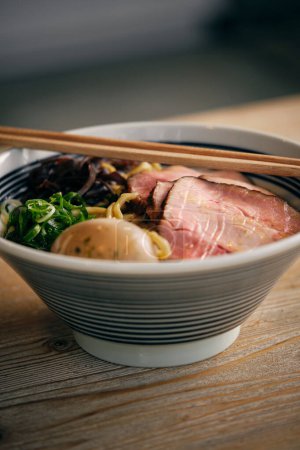 Téléchargez les photos : Bol de délicieuse soupe ramen japonaise avec des morceaux de viande de porc avec des baguettes placées sur une table en bois dans le restaurant - en image libre de droit