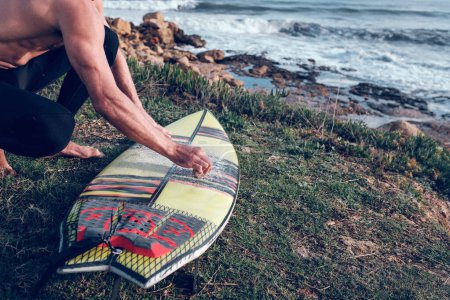 Photo for From above of crop naked torso surfer man covering surfboard with wax sitting on squat on grassy coast - Royalty Free Image