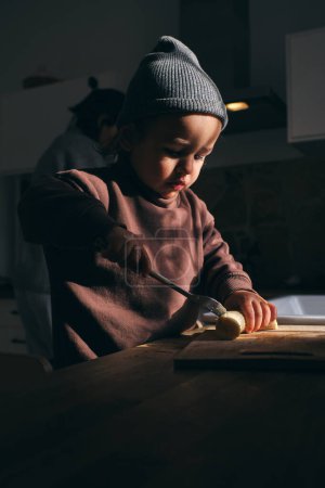 Photo for Focused little boy in casual clothes and knitted hat cutting ripe banana with knife on cutting table while standing near table in kitchen - Royalty Free Image