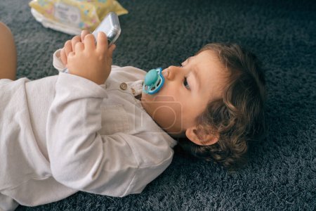 Photo for From above of curious little boy with pacifier lying on floor and looking at screen of mobile phone in living room at home - Royalty Free Image