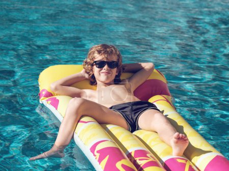 Hoher Winkel von positivem blondem Kind in Shorts und Sonnenbrille, das die Hände hinter dem Kopf hält und lächelt, während es sich auf einem aufblasbaren Schwimmer im Schwimmbad auf Mallorca, Spanien, entspannt 