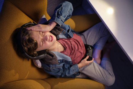 Photo for From above of curly haired preteen kid in denim jacket sitting in armchair with crossed legs and joystick while doing facepalm gesture because of losing game - Royalty Free Image