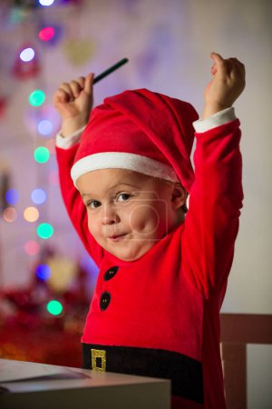 Foto de Encantador niño en traje de Santa sentado con los brazos levantados en la mesa mientras sostiene el lápiz y mirando a la cámara en la habitación decorada - Imagen libre de derechos