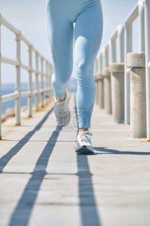 Photo for Crop unrecognizable female athlete in sportswear and sneakers jogging on bridge with shadows during workout in sunlight - Royalty Free Image