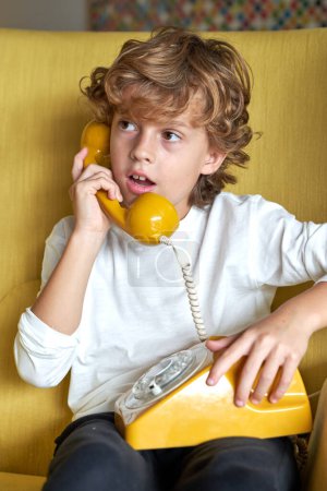 Photo for Thoughtful kid in casual wear speaking on vintage landline telephone while sitting in armchair during conversation in light living room - Royalty Free Image